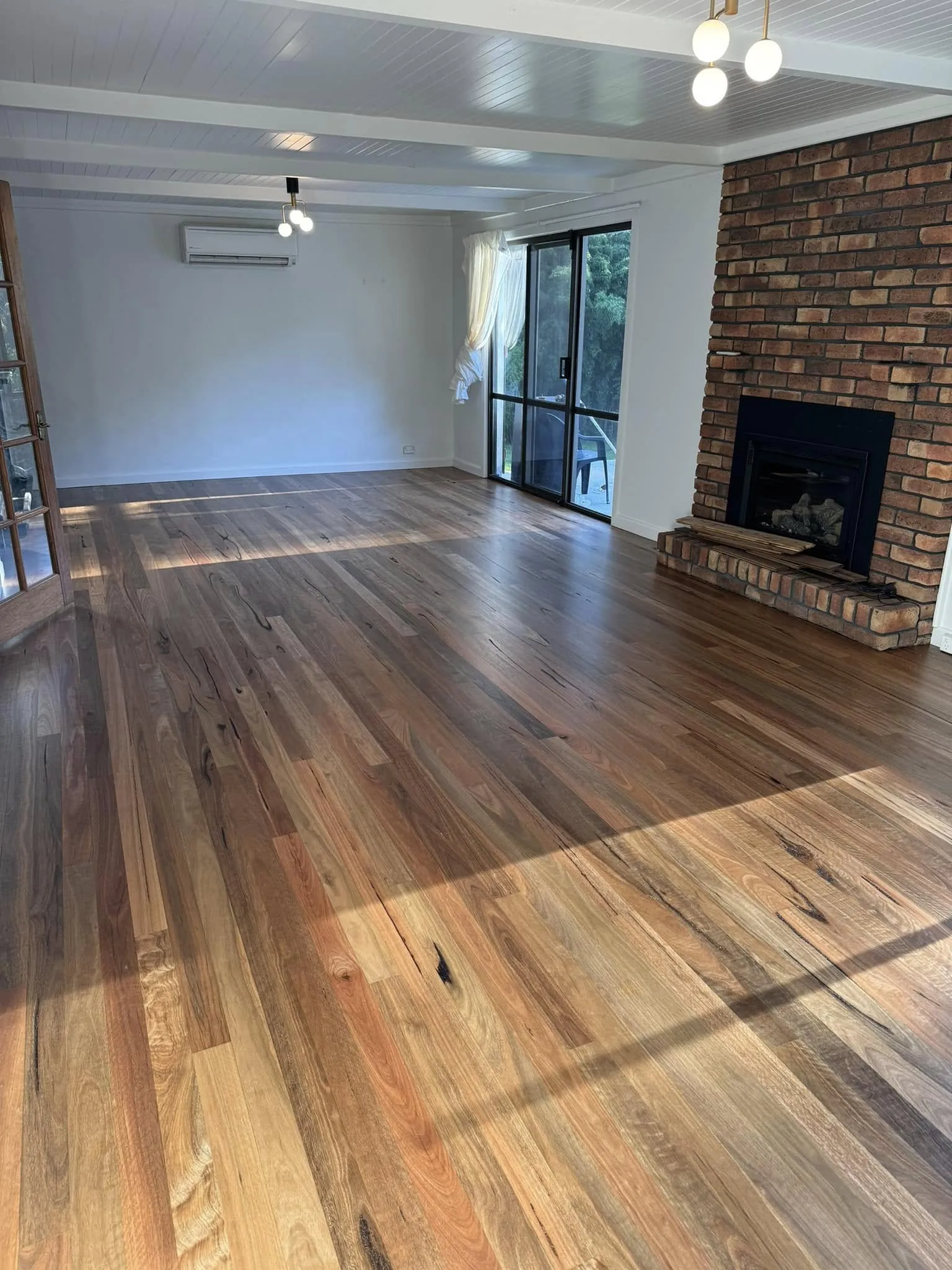 A restored hardwood floor running through a lounge room to a brick fireplace and glass doors